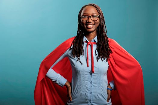 Proud and brave superhero woman wearing mighty hero red cape standing on blue background. Positive and optimistic tough looking young person wearing cloak while smiling heartily and looking at camera