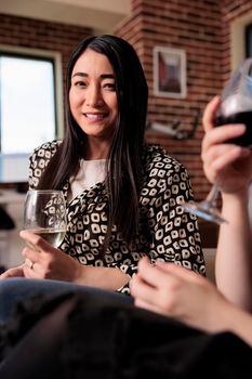 Smiling heartily young adult woman drinking wine at birthday event party while looking at camera. Beautiful girl enjoying hanging out with friends at home while celebrating important life moment.