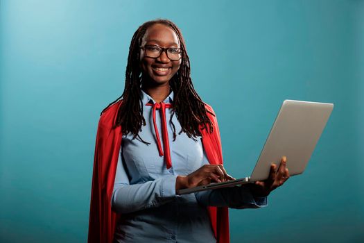 Happy joyful superhero woman wearing mighty hero red cloak while standing on blue background. Portrait of confident and positive african american justice defender browsing internet on modern laptop.