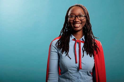 Portrait of african american young adult superhero woman wearing red cape on blue background while smiling at camera. Studio shot of beautiful and joyful justice defender posing cheerful.