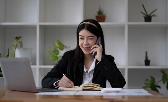 Sharing good business news. Attractive young woman talking on the mobile phone and smiling while sitting at her working place in office and looking at laptop.