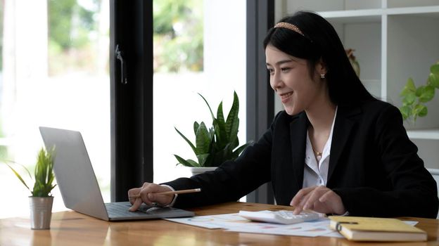 Beautiful Asian businesswoman analyzes charts using laptop calculator at the office..