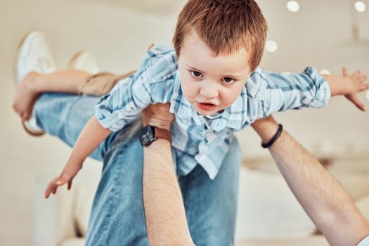 Adorable little caucasian boy lift into the air by his father so he can pretend to fly like a plane or superhero with arms out while relaxing at home. Loving dad playing and bonding with carefree kid.