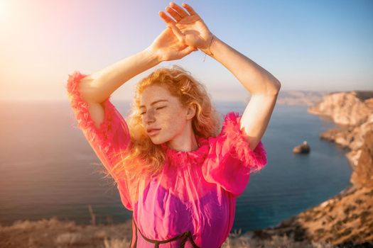 Close up shot of beautiful young caucasian woman with curly blond hair and freckles looking at camera and smiling. Cute woman portrait in a pink long dress posing on a volcanic rock high above the sea