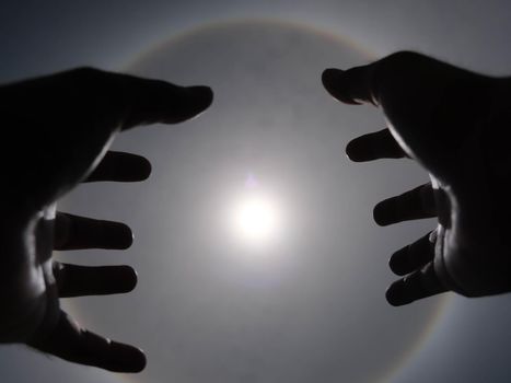 Beautiful photograph of the sun with a circular rainbow surrounded by a bright sky and white clouds with shadows of hands reaching out. Phenomenon, sun halo.