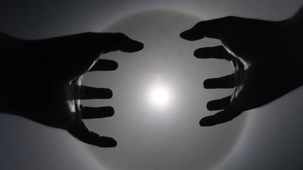 Beautiful photograph of the sun with a circular rainbow surrounded by a bright sky and white clouds with shadows of hands reaching out. Phenomenon, sun halo.