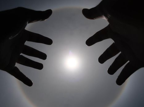 Beautiful photograph of the sun with a circular rainbow surrounded by a bright sky and white clouds with shadows of hands reaching out. Phenomenon, sun halo.