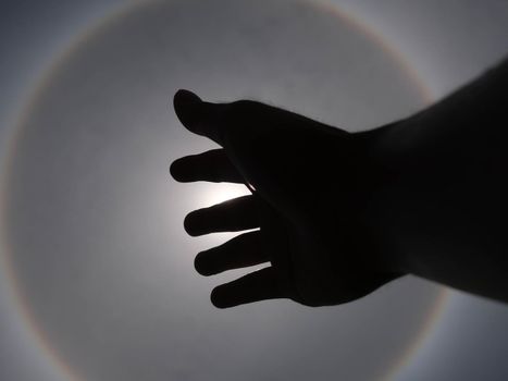 Beautiful photograph of the sun with a circular rainbow surrounded by a bright sky and white clouds with shadows of hands reaching out. Phenomenon, sun halo.