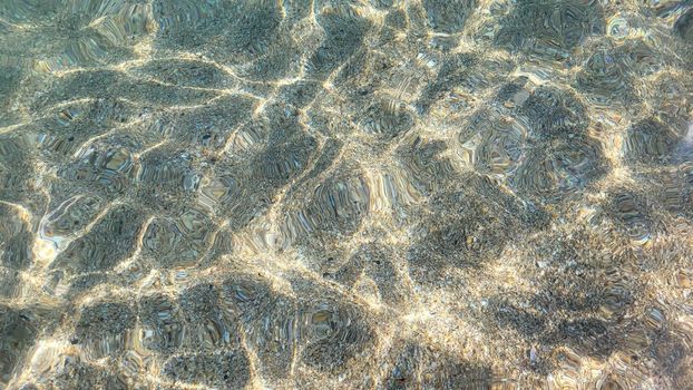 Sea sandy bottom is visible through clear turquoise sea water. Sun glare on sea surface. Beautiful abstract background. Copy space. Selective focus.