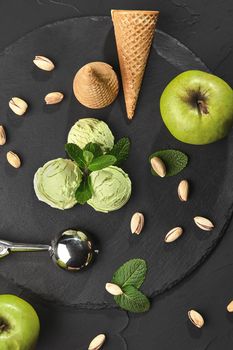 Top view of an appetizing pistachio ice cream decorated with mint, waffle cones with scattered pistachios and green apples are nearby, served with a metal scoop on a dark slate over a black background. Close-up.