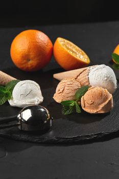 Side view of a stone slate tray with an excellent creamy and orange ice cream set decorated with mint, and waffle cones on a dark table over a black background. Metal scoop is laying nearby. Summer coolness of ice cream and sorbet.