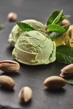 Close-up shot of a tasty pistachio ice cream decorated with mint, scattered pistachios are nearby, served on a stone slate over a black background.
