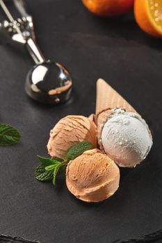Stone slate tray with an appetizing creamy and orange ice cream set decorated with fresh mint, and classic waffle cones on a dark table over a black background. Metal scoop is laying nearby. Summer coolness of ice cream and sorbet. Close-up shot.