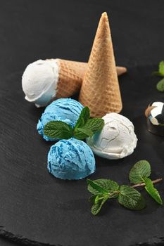 Top view of fantastic creamy and blueberry ice cream decorated with mint, and waffle cones are served on a stone slate standing on a dark table over a black background. Metal scoop is laying nearby. Close-up shot.