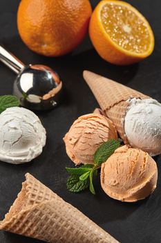 Stone slate tray with a fresh creamy and orange ice cream set decorated with mint, and classic waffle cones on a dark table over a black background. Metal scoop is laying nearby. Summer coolness of ice cream and sorbet. Close-up shot.