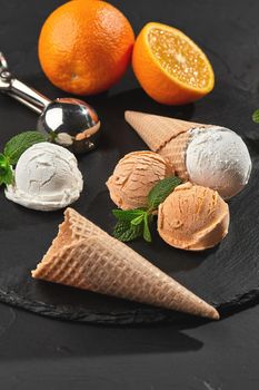 Close-up shot of a stone slate tray with a homemade creamy and orange ice cream set decorated with fresh mint, and classic waffle cones on a dark table over a black background. Metal scoop is laying nearby. Summer coolness of ice cream and sorbet.