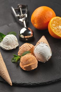 Close-up shot of a stone slate tray with an enjoyable creamy and orange ice cream set decorated with fresh mint, and waffle cones on a dark table over a black background. Metal scoop is laying nearby. Summer coolness of ice cream and sorbet.