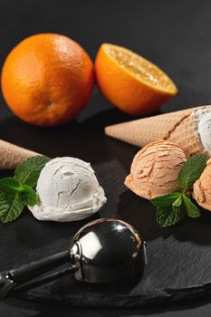 Side view of a stone slate tray with a creamy and orange ice cream set decorated with fresh mint, and classic waffle cones on a dark table over a black background. Metal scoop is laying nearby. Summer coolness of ice cream and sorbet. Close-up shot.