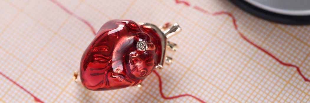 Close-up of red glass miniature heart toy lay over heart cardiogram result. Examination of cardiovascular system. Medicine, cardiology, healthcare concept