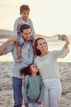 Happy cheerful mixed race family smiling for a selfie spending time at the beach together. Hispanic mother smiling taking a photo with her children and husband with her cellphone bonding on vacation.