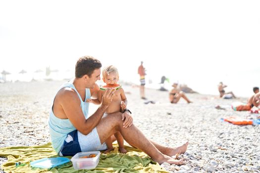 Smiling dad feeding little girl with slice of watermelon on beach. High quality photo
