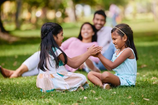 Two little girls playing a clapping game while sitting in the park on a sunny day. Parent watching their daughters get along. Small sibling sister friends playing together on the grass.