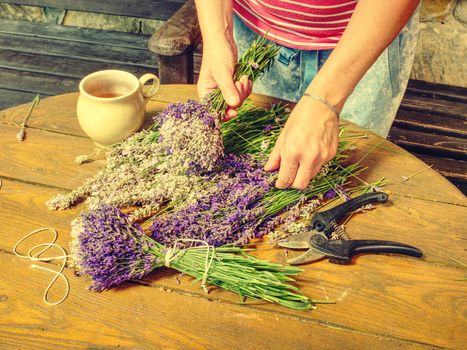 Skilful hands of girl putting fresh blossoms of  lavender into wonderfully scented bouquet. Flowers and  garden shears on wooden table workplace 