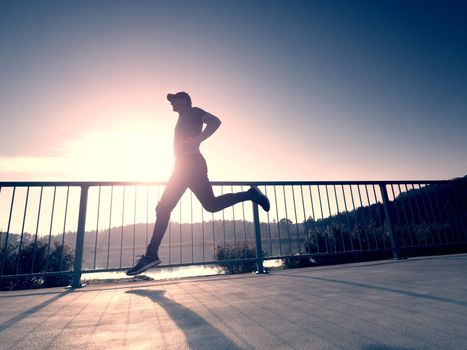 Attractive wiry man jogging against to sun. Regular morning training at sea. Young sportsman jogging