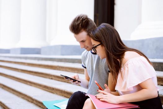 Man showing girl interesting content on his mobile phone while resting on stairs of university campus