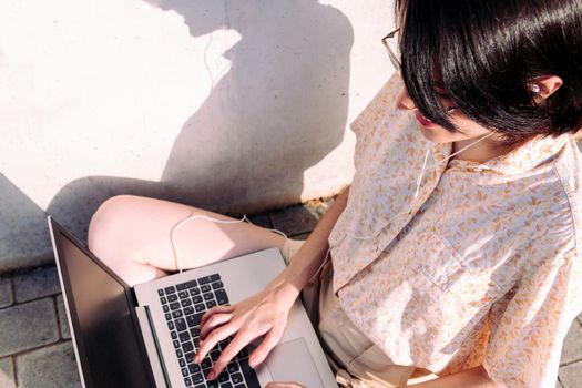 top view of an unrecognizable young woman with earphones working with a laptop sitting next to a gray wall, technology concept and blogging lifestyle, text copy space