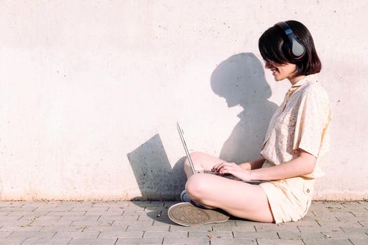 profile of a young woman with headphones working with a laptop sitting next to a gray wall, technology concept and urban lifestyle, text copy space
