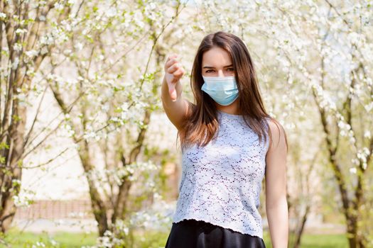 Girl stanting in blooming park showing thumbs down, to show her irritation and dislike to constant wearing medical mask to prevent spread of coronavirus