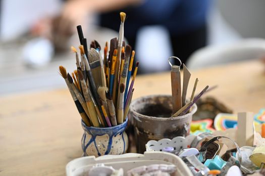 Ceramics making equipment with paint brushes, pottery shaping tools on wooden table in creative studio.