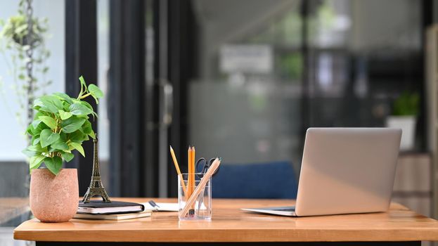 Wooden office desk with laptop computer, notebooks, pencil holder and potted plant.