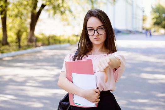 Female student showing big finger down showing gesture of bad idea standing with learning materials in the park after classes