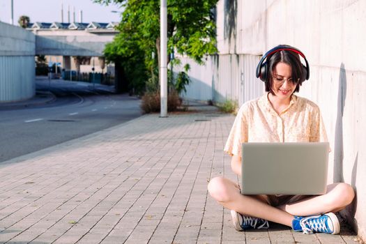 front view of a young woman with headphones working with a computer sitting next to a gray wall, technology concept and urban lifestyle, text copy space