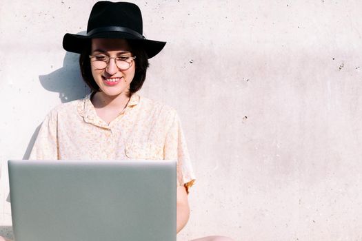 young woman with hat working with computer sitting against a grey wall, concept of digital nomad and blogging lifestyle , copy space for text