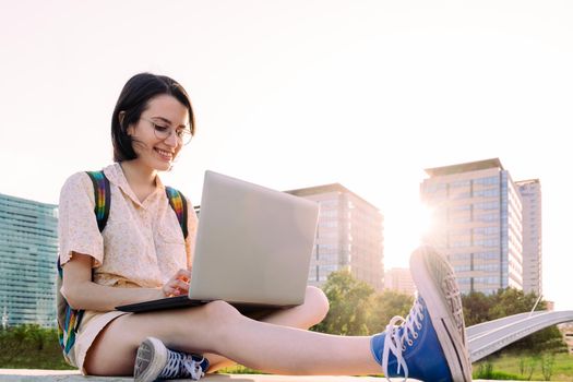 happy young traveler woman with backpack sitting in the city typing on her laptop, concept of technology, youth and digital nomad lifestyle, copy space for text