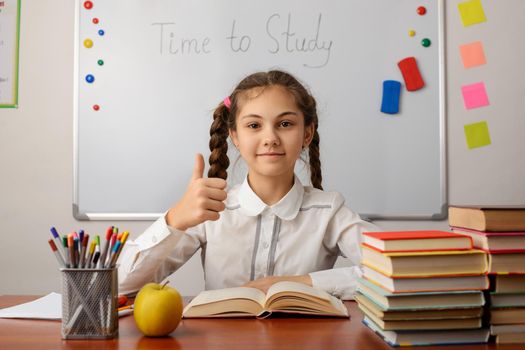 Cheerful pupil showing thumbs up gesture, studying at the classroom, reading a book