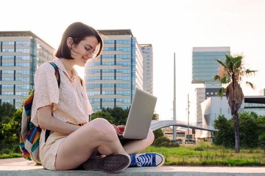 young traveler woman with backpack sitting in the city typing on her computer, concept of technology, youth and digital nomad lifestyle