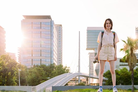 smiling young traveler woman with backpack and camera standing in front of the city looking to camera, concept of youth and digital nomad lifestyle, copy space for text