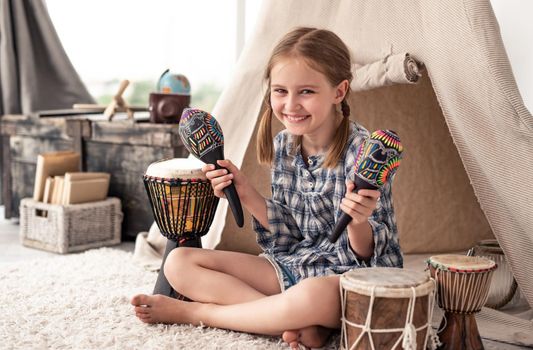 Smiling little girl shaking music maracas sitting in room with djembe drums and wigwam