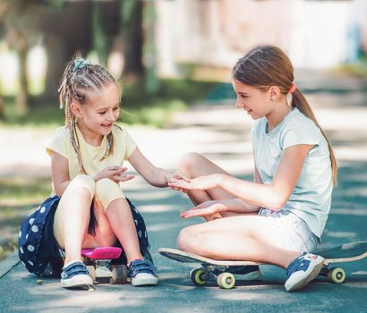 Smiling little kids girls sitting on skateboards in the park and showing manicure