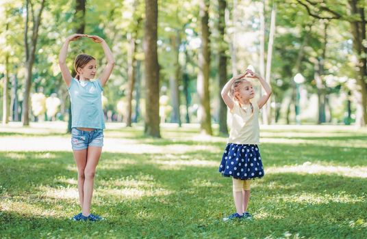 Beautiful girl with her little sister making ballet training together in park outside in summertime