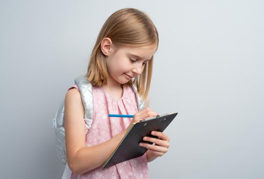 Little girl taking notes with pencil standing against gray wall