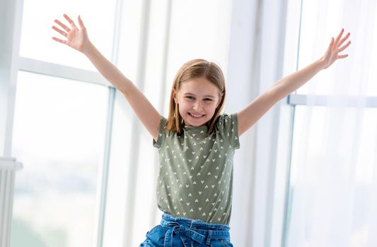 Portrait of preteen beautiful girl posing in light room with hands up and smiling. Female kid schoolgirl looking at the camera indoors