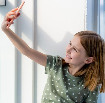 Cute preteen girl sitting on windowsill with smartphone and making selfie with daylight from window. Beautiful female kid schoolgirl posing with cell phone at home