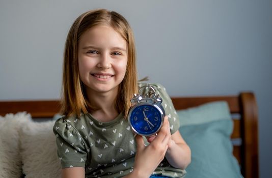 Portrait of preteen beautiful blond hair girl with blue clock in bedroom. Female kid schoolgirl sitting in the bed and looking at the camera smiling