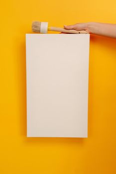 Hand holding white canvas with copy space and wooden paint brush tool on yellow background. Empty sheet banner with place for text. Concept of design, art creativity