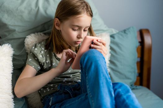 Pretty preteen girl sitting in the bed and looking at smartphone at home in the bedroom. Beautiful kid with cell phone indoors